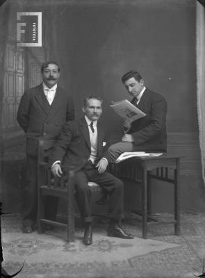 Carlos Bustos leyendo sentado en mesita, Olivera Córdoba parado y Rück Uriburu sentado en el medio